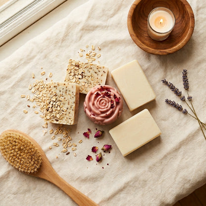 2 smooth pale ivory Rice Milk bars, 2 cream-colored bars with visible oat flecks (Almond Oats), and 1 soft rose-pink, rose-shaped Yoni bar finished with a few dried rose petals