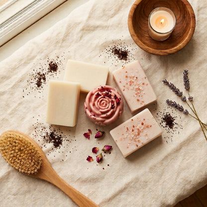 2 smooth ivory-white Goat's Milk bars, 2 soft blush-white bars with visible pink Himalayan salt crystals (Salted Spa), and 1 soft rose-pink, rose-shaped Yoni bar finished with a few dried rose petals