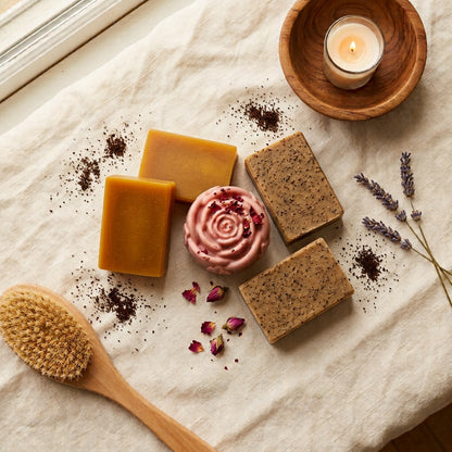 2 smooth golden-amber Turmeric bars, 2 tan-brown bars with visible flecks of coffee grounds (Coffee Mint), and 1 soft rose-pink, rose-shaped Yoni bar finished with a few dried rose petals
