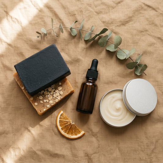 A styled flat-lay of natural skincare products on a textured tan fabric background, including a black soap bar, an amber dropper bottle, a silver tin of cream, oats, a dried orange slice, and eucalyptus leaves.