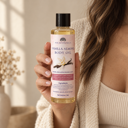 A woman in a cream knit sweater holding the 8 oz Vanilla Almond Pure Body Oil bottle in one hand, in soft window light — PureButterMelts